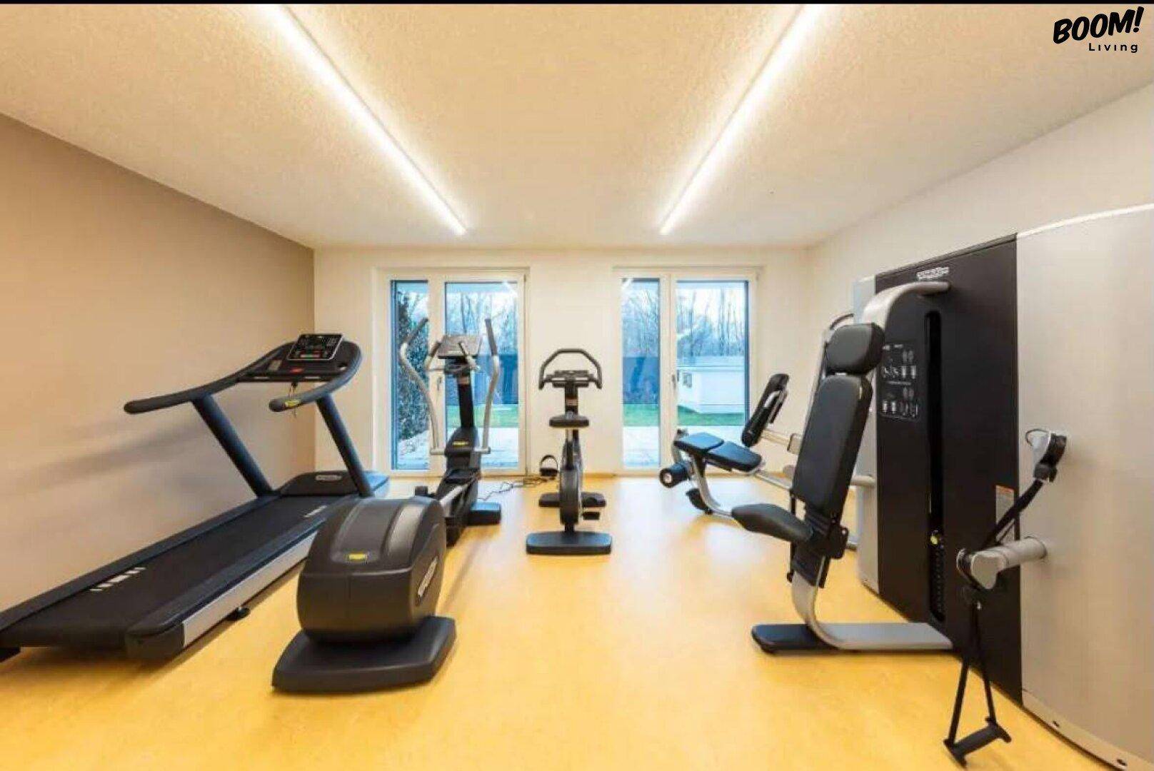 Moderner Fitnessraum mit verschiedenen Trainingsgeräten und großen Fenstern für Tageslicht.