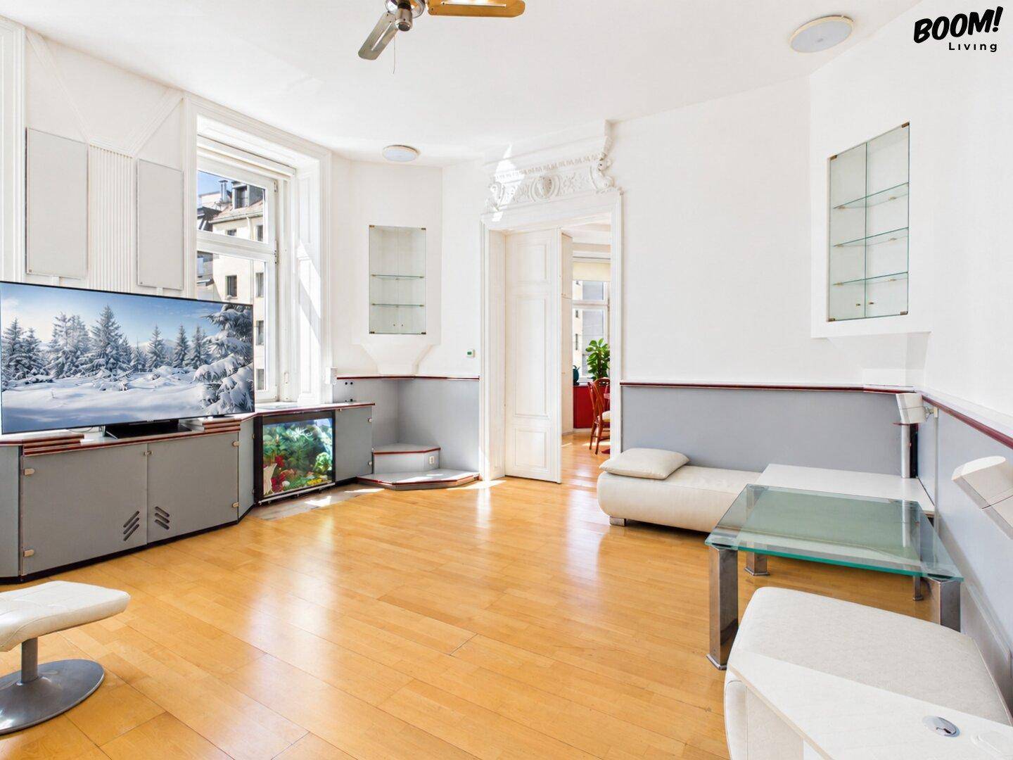 Helles Wohnzimmer mit Holzboden, großem Fernseher und minimalistischer Einrichtung, durch eine Tür mit einem weiteren Raum verbunden.