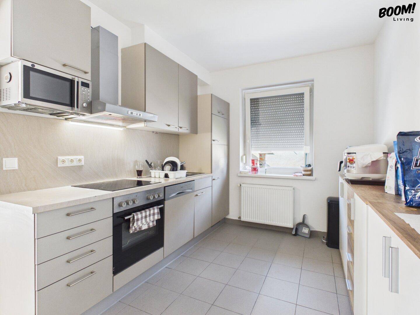 Moderne Küche mit grauen Schränken, Einbaugeräten und Fenster mit Rollladen.