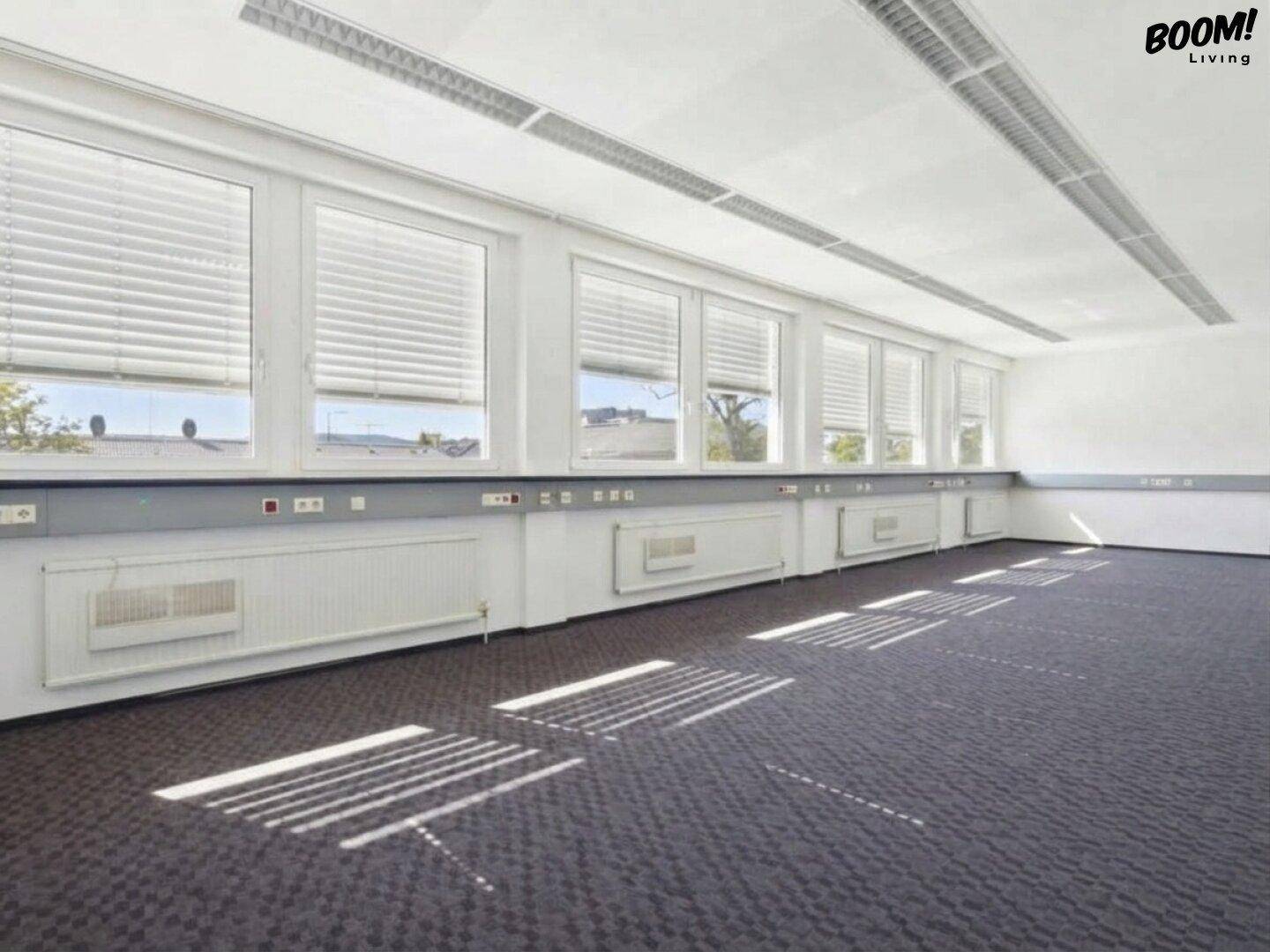 Geräumiges, leeres Büro mit vielen Fenstern, Heizkörpern und Teppichboden, bereit zur Einrichtung.
