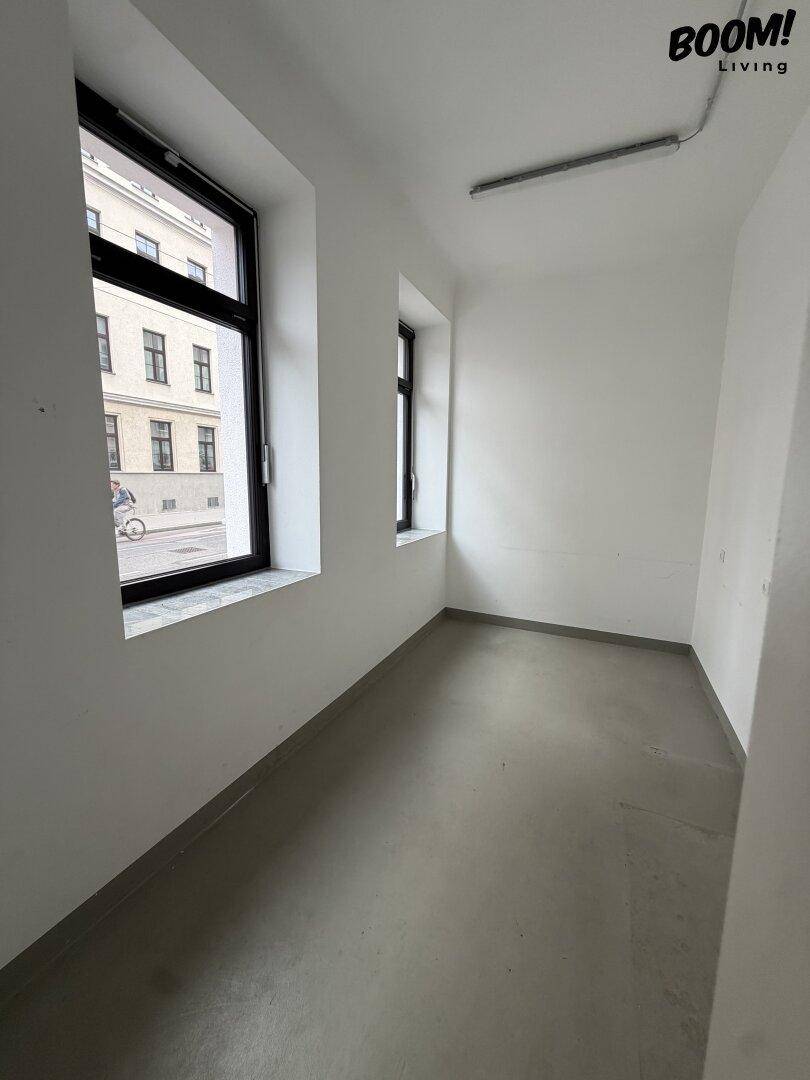 Schmaler Büroraum mit Betonboden, weißen Wänden und Fenstern mit Blick auf die Straße.
