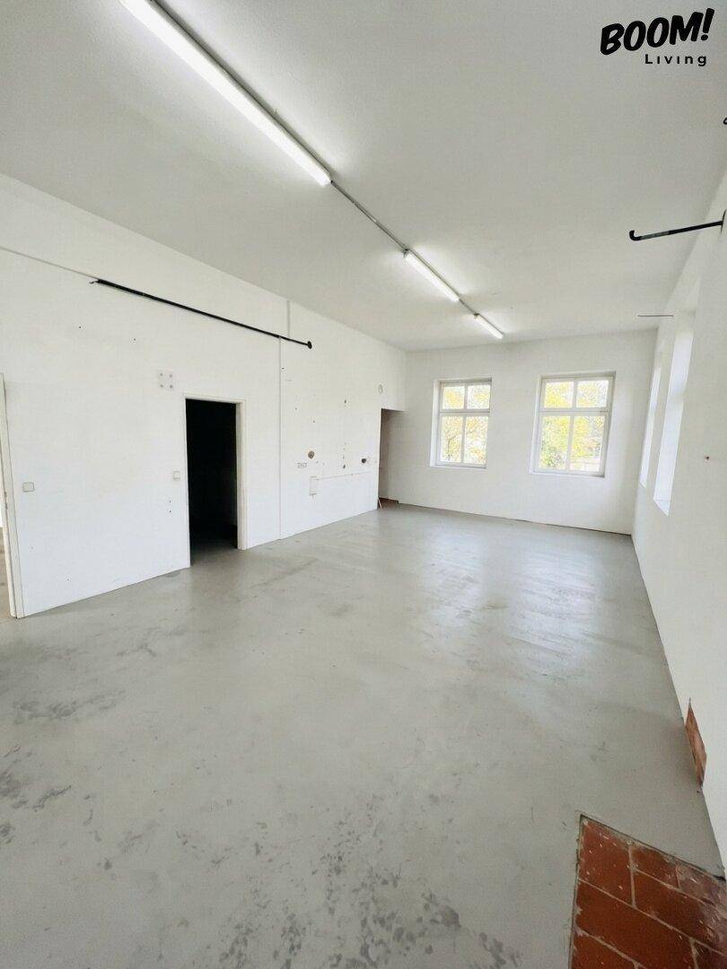 Geräumiger Büroraum mit Betonboden, weißen Wänden und mehreren Fenstern, die viel Licht hereinlassen.