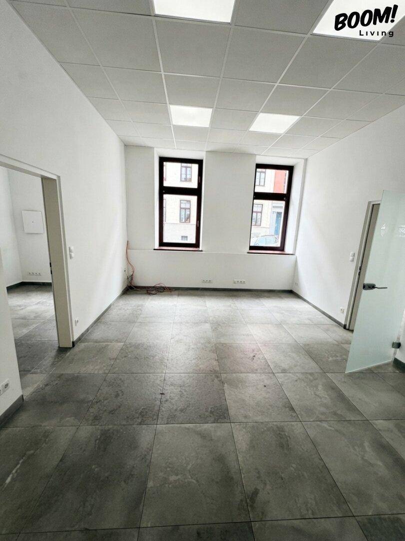 Leerer Büroraum mit zwei Fenstern, grauen Fliesen und einer Decke mit integrierten Leuchten.