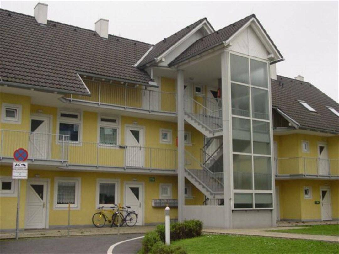 Gelbe Gebäudefassade mit Außentreppe und verglastem Aufzug für barrierefreien Zugang.