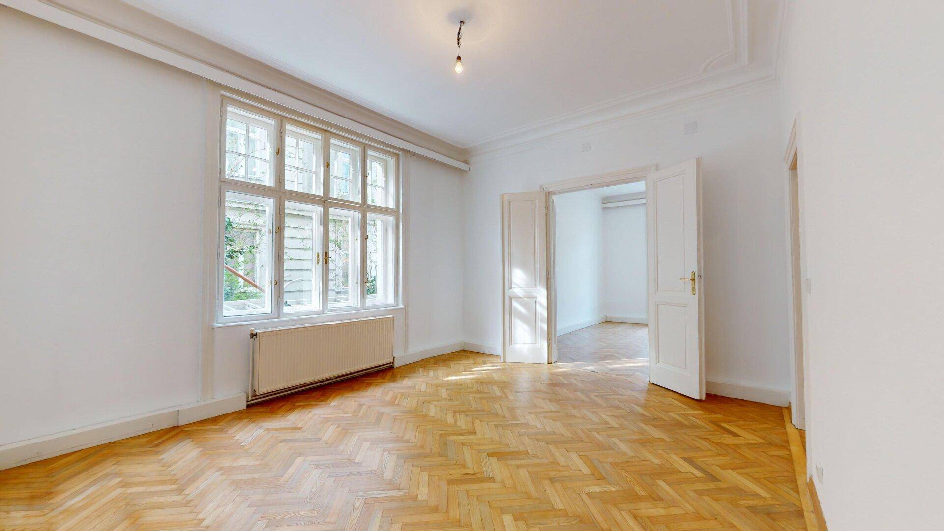 Geräumiges Zimmer mit klassischem Fischgrätparkett und viel Tageslicht durch große Fenster.