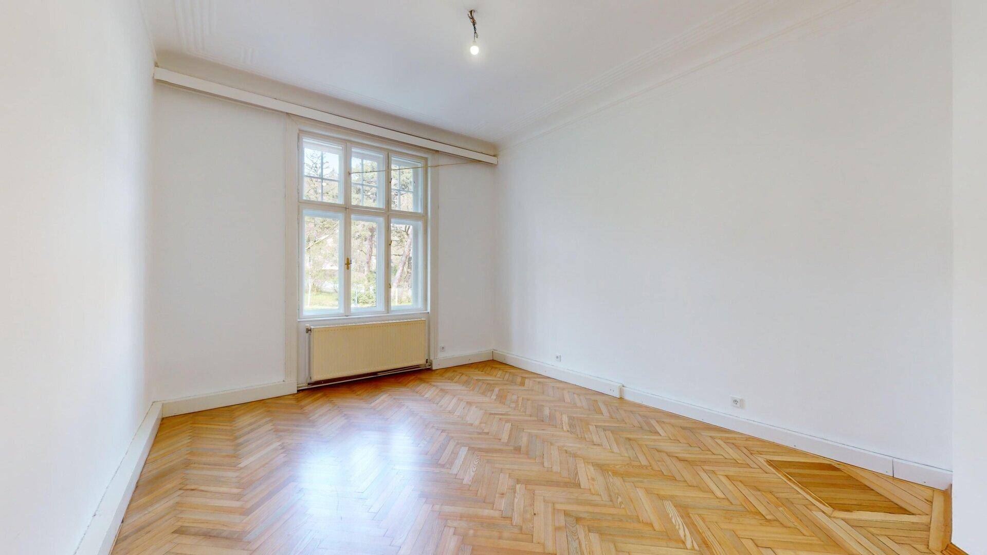 Heller Raum mit klassischem Fischgrätparkett und einem großen Fenster mit Blick ins Grüne.