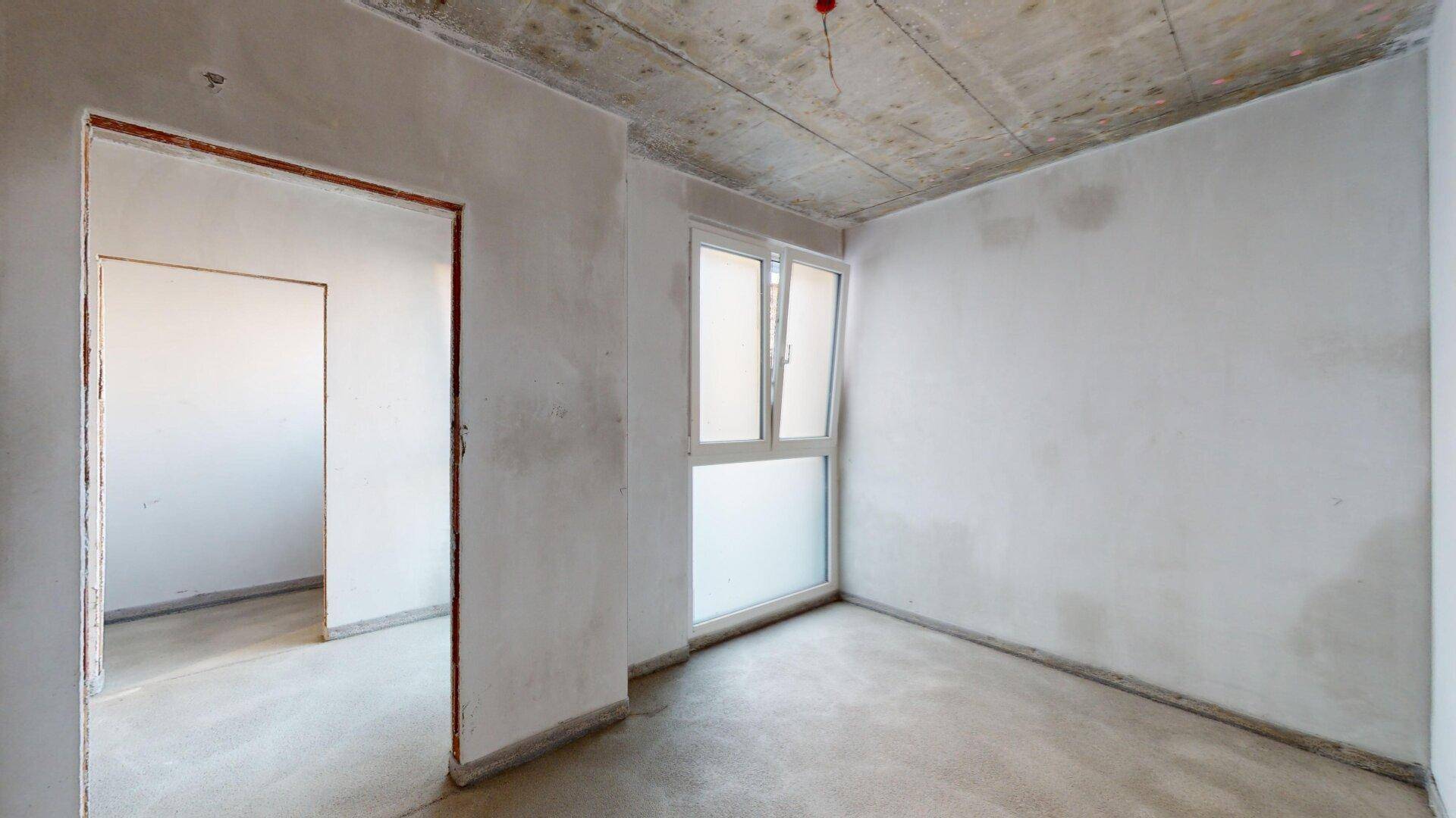 Rohbau-Raum mit Betonboden und -decke sowie einem Fenster für Tageslicht.