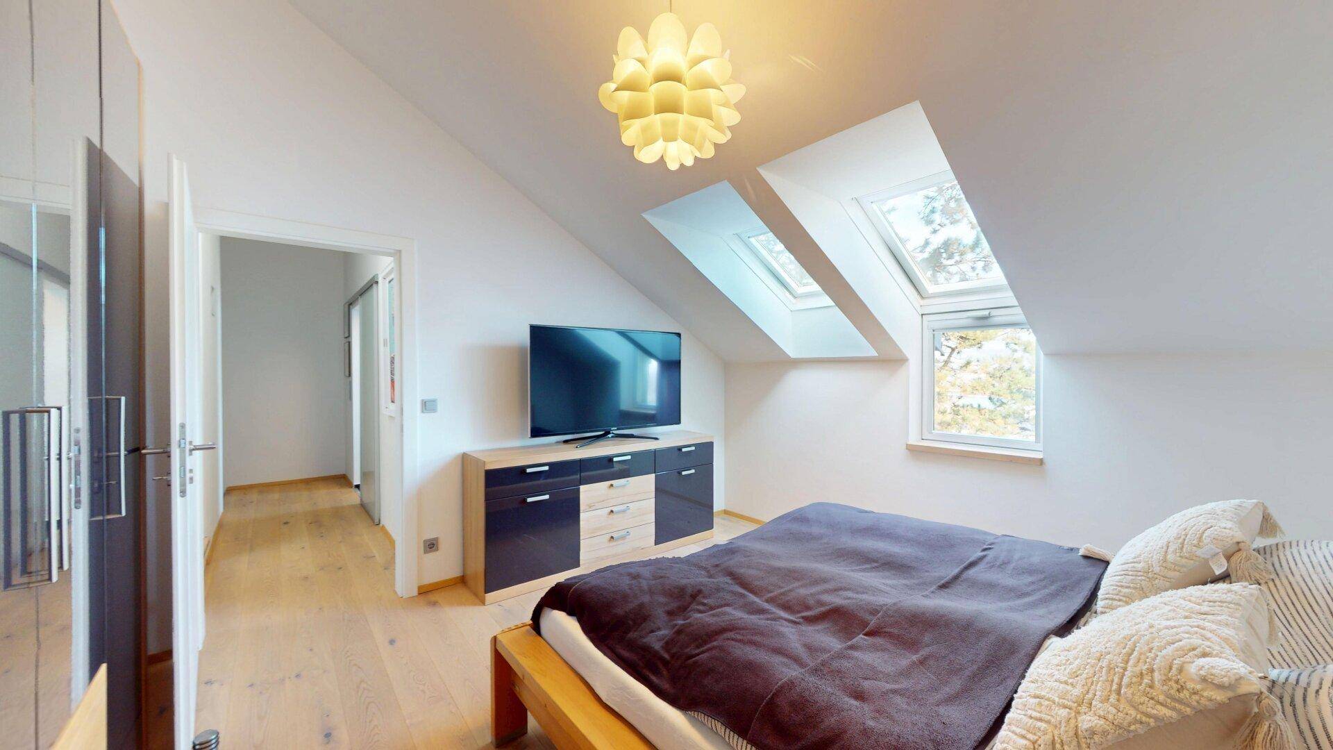 Helles Schlafzimmer im Dachgeschoss mit Doppelbett, Fernseher und großzügigen Dachfenstern.