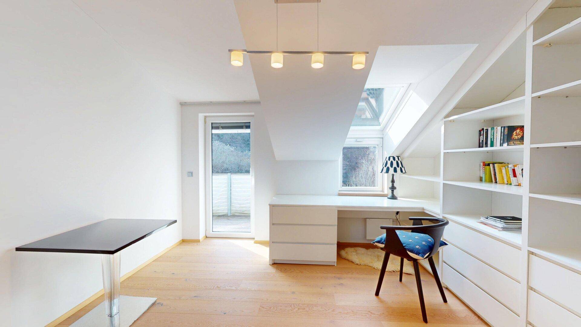 Helles Arbeitszimmer im Dachgeschoss mit Schreibtisch, Regalwand und Zugang zu einem kleinen Balkon.