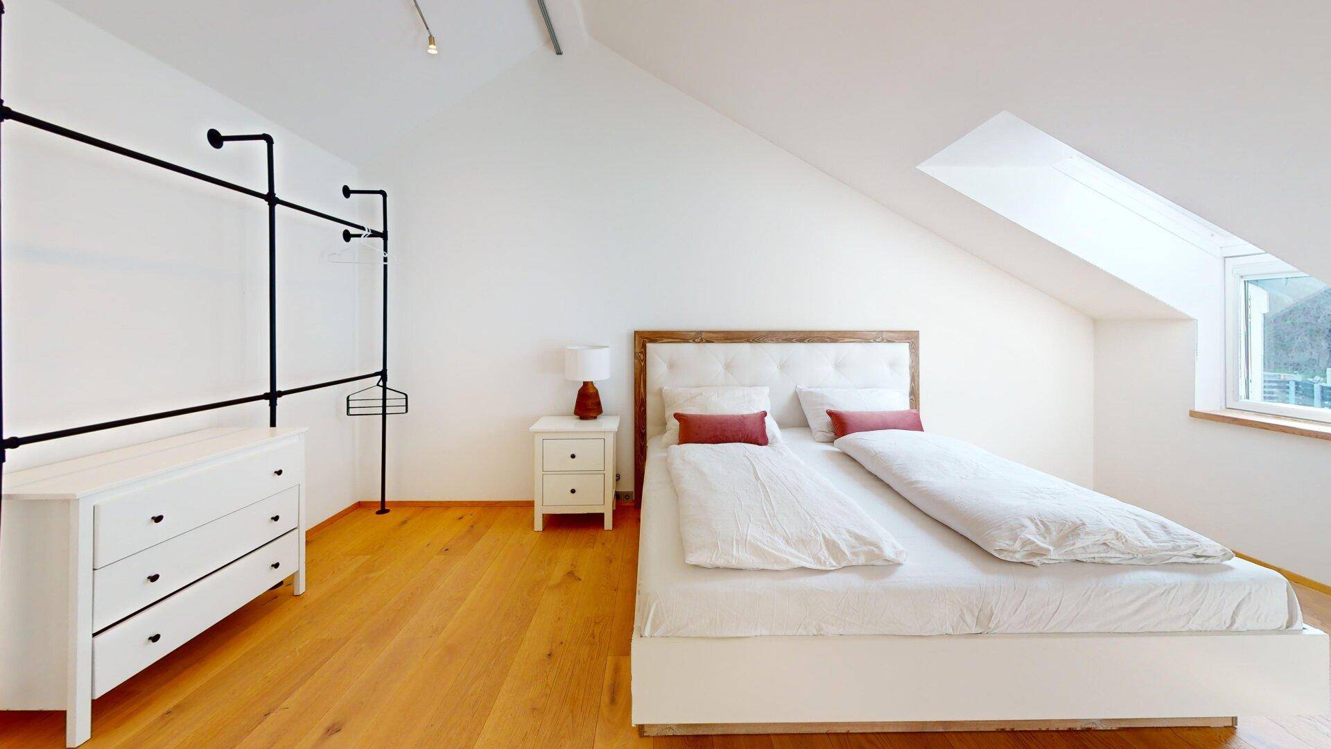 Helles Schlafzimmer mit Holzboden, offenem Kleiderschrank und Dachschräge mit Fenster.