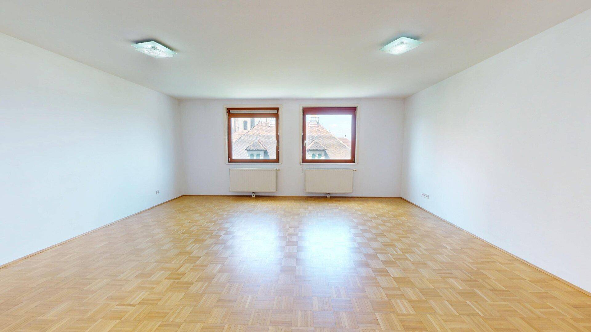 Heller, leerer Raum mit Parkettboden und zwei Fenstern mit Blick auf Dächer.