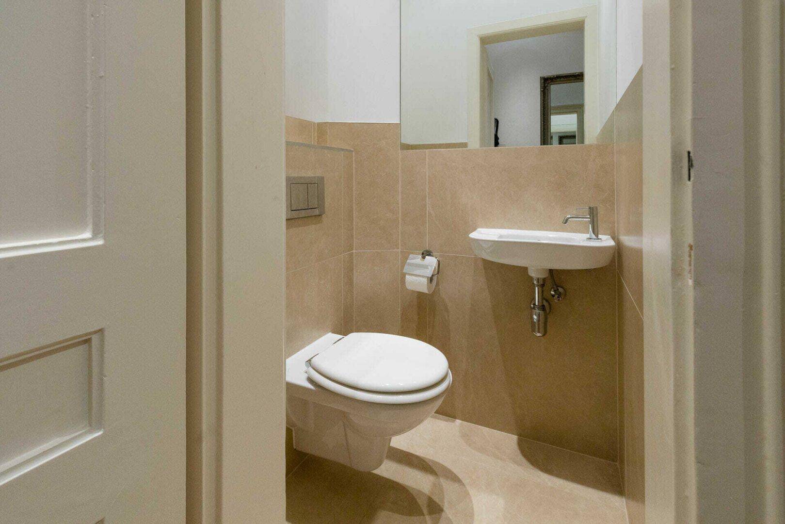 Kompaktes Gäste-WC mit Wand-WC, kleinem Waschbecken und beigefarbenen Fliesen.