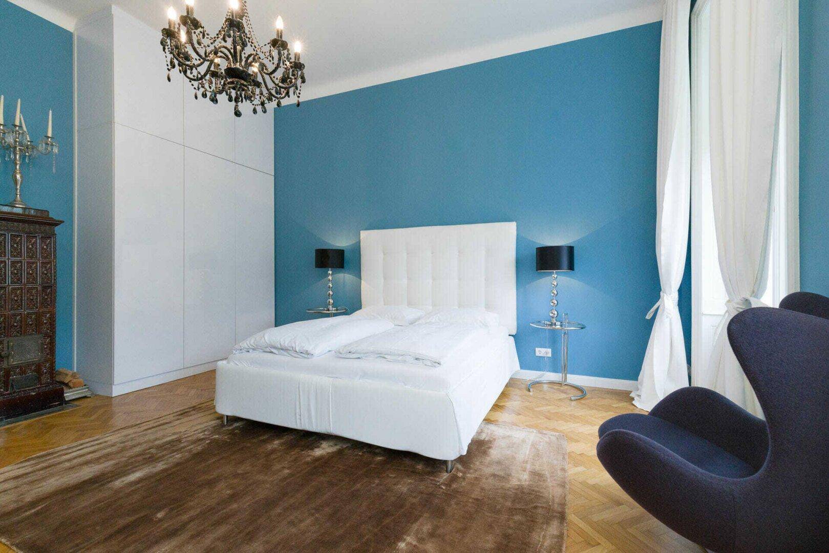 Elegantes Schlafzimmer mit blauer Akzentwand, Fischgrätparkett und einem opulenten schwarzen Kronleuchter.