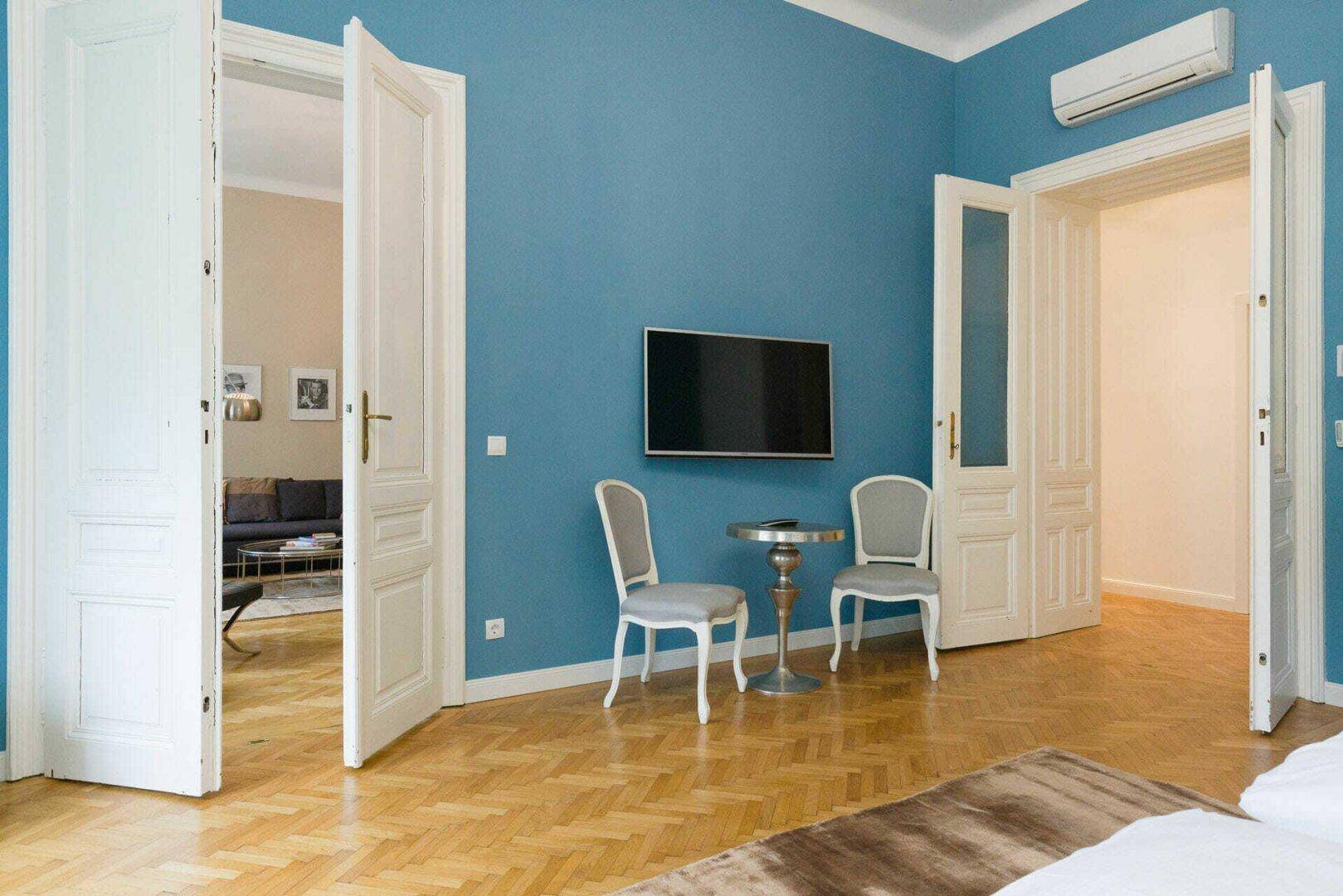 Schlafzimmer mit blauer Wand, Fernseher und Zugang zu einem weiteren Raum durch Flügeltüren.