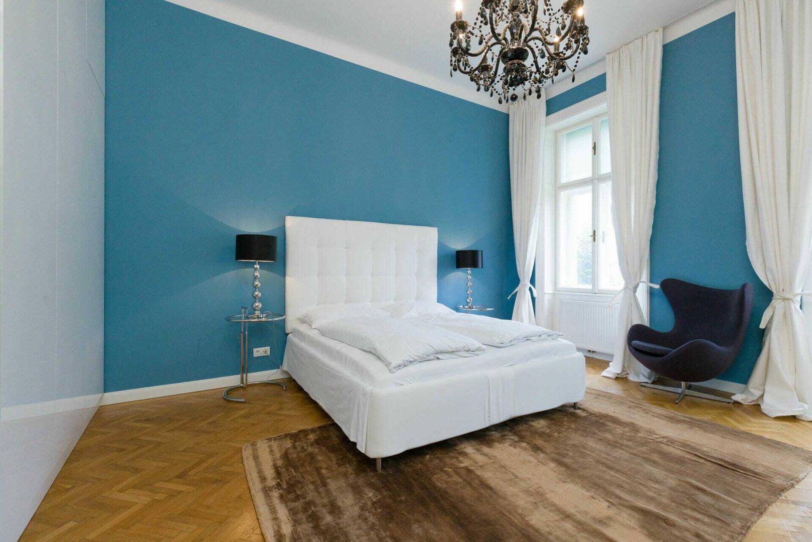 Helles Schlafzimmer mit blauer Wand, großem Fenster und klassischem Fischgrätparkettboden.
