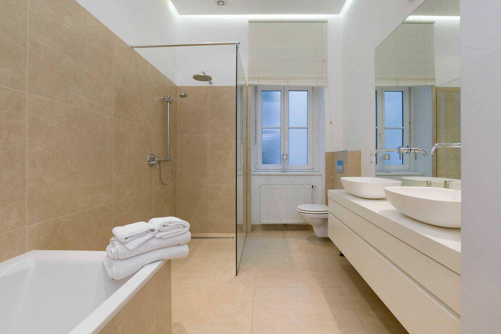 Helles Badezimmer mit Badewanne, Dusche, Doppelwaschbecken und einem Fenster für Tageslicht.