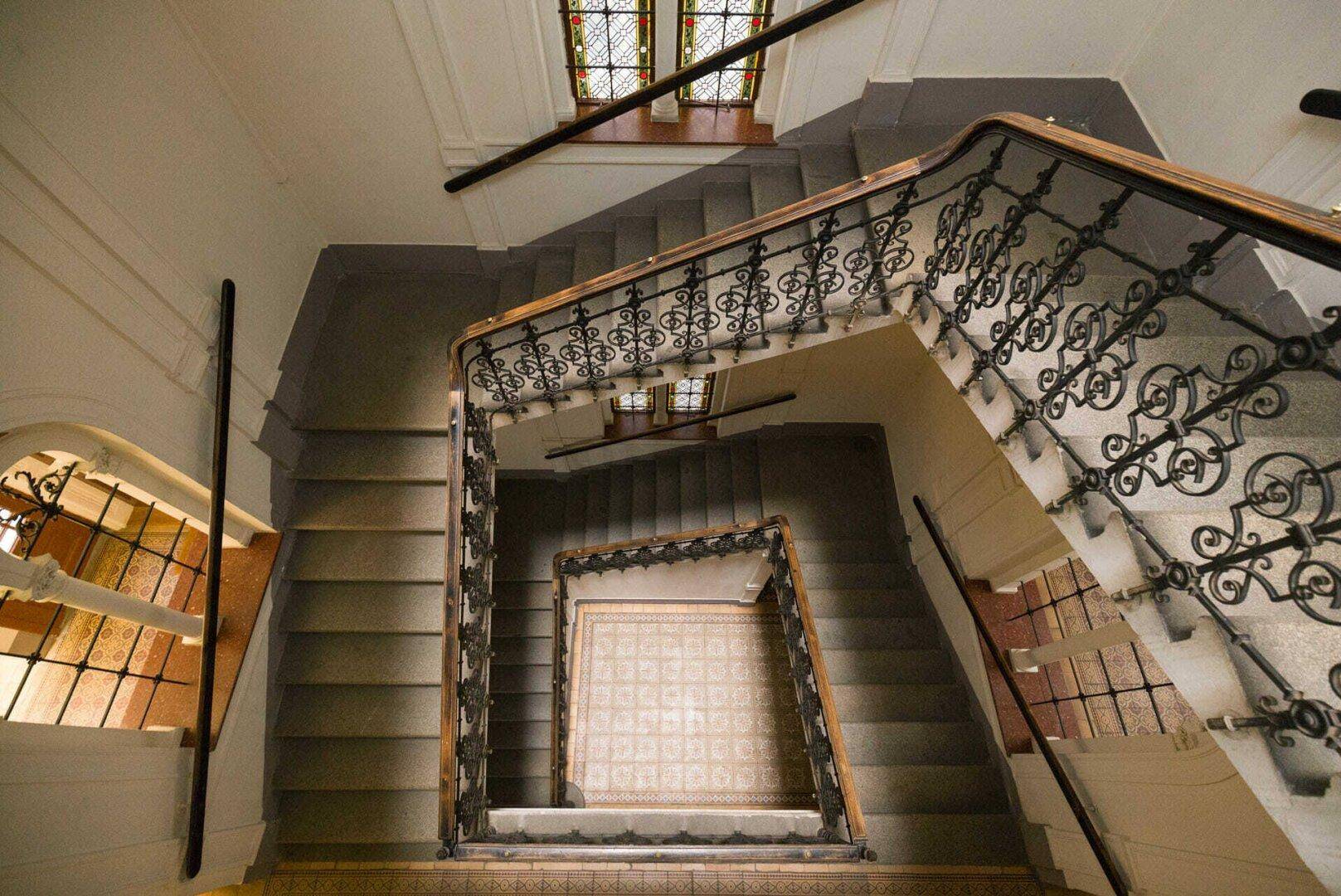 Historisches Treppenhaus von oben gesehen mit kunstvollen Geländern und gemusterten Fliesen.