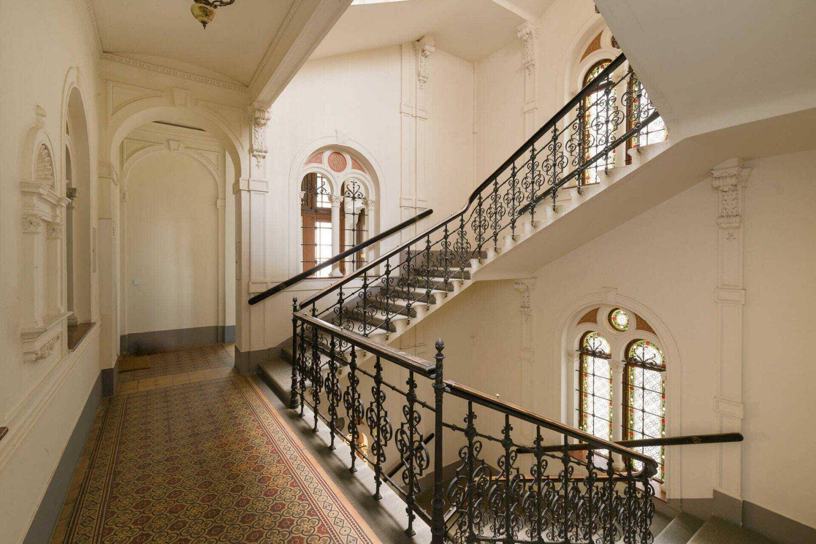 Historisches Treppenhaus mit kunstvollen Geländern, Stuckverzierungen und großen Fenstern.