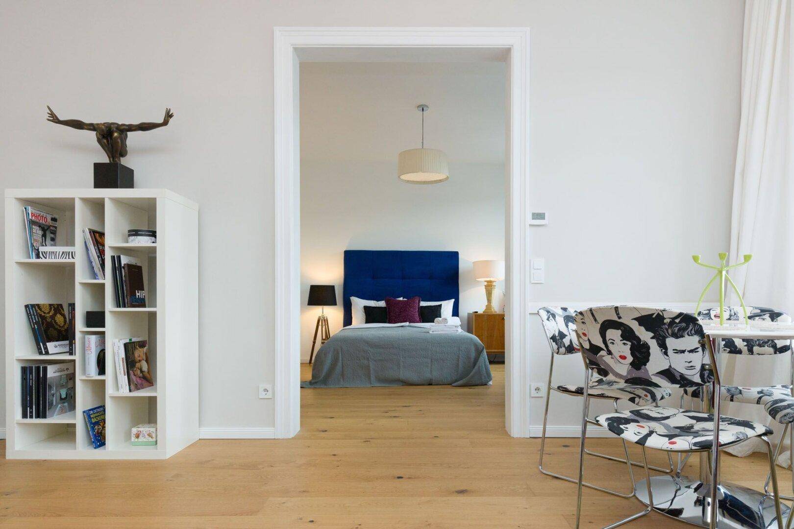Offener Übergang vom Wohnbereich mit Bücherregal zum Schlafzimmer mit blauem Kopfteil.