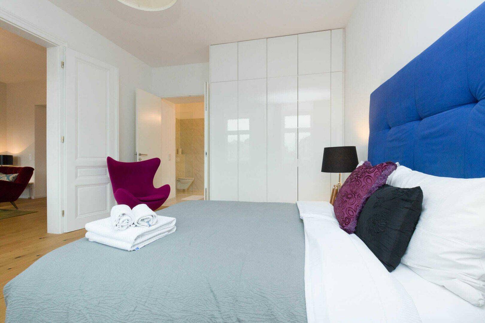Schlafzimmer mit blauem Kopfteil, weißen Einbauschränken und Zugang zum Badezimmer.