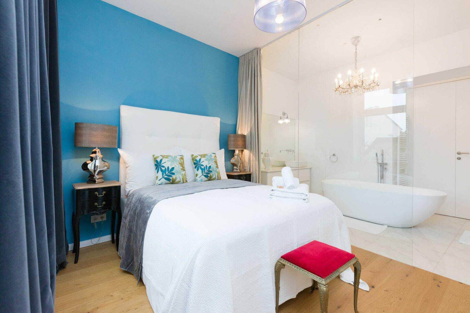 Schlafzimmer mit blauer Akzentwand, Holzboden und offener Verbindung zum modernen Badezimmer.
