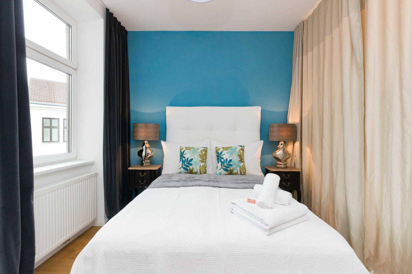 Gemütliches Schlafzimmer mit blauer Akzentwand, großem Fenster und Nachttischen.