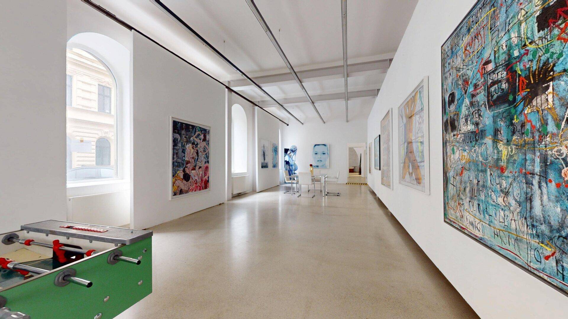 Heller Ausstellungsraum mit Kunstwerken an den Wänden, hohen Fenstern und einem Tischfußballtisch.