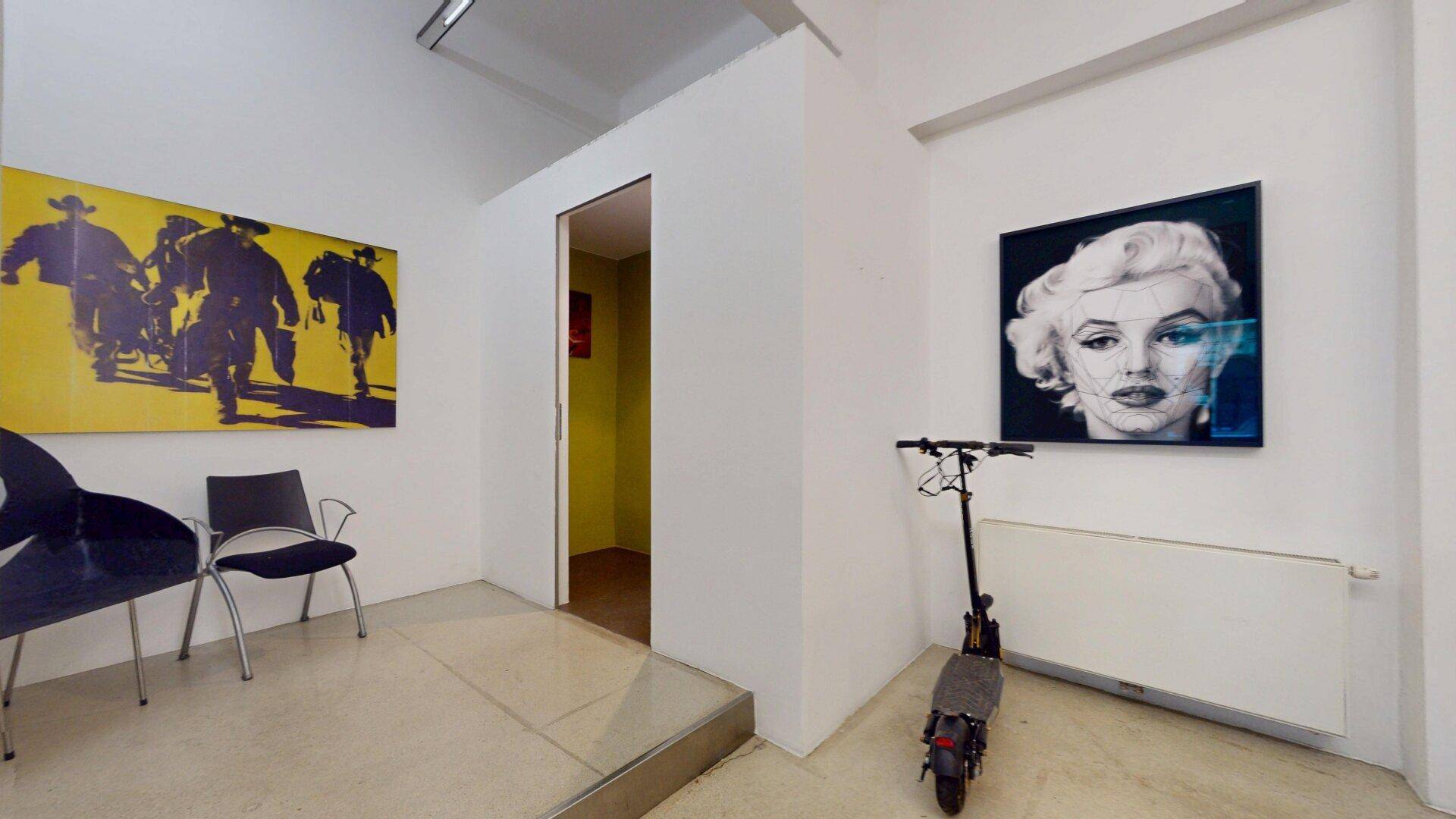 Ecke eines Ausstellungsraums mit Kunstwerken, Sitzgelegenheiten und einem Durchgang zu einem weiteren Raum.