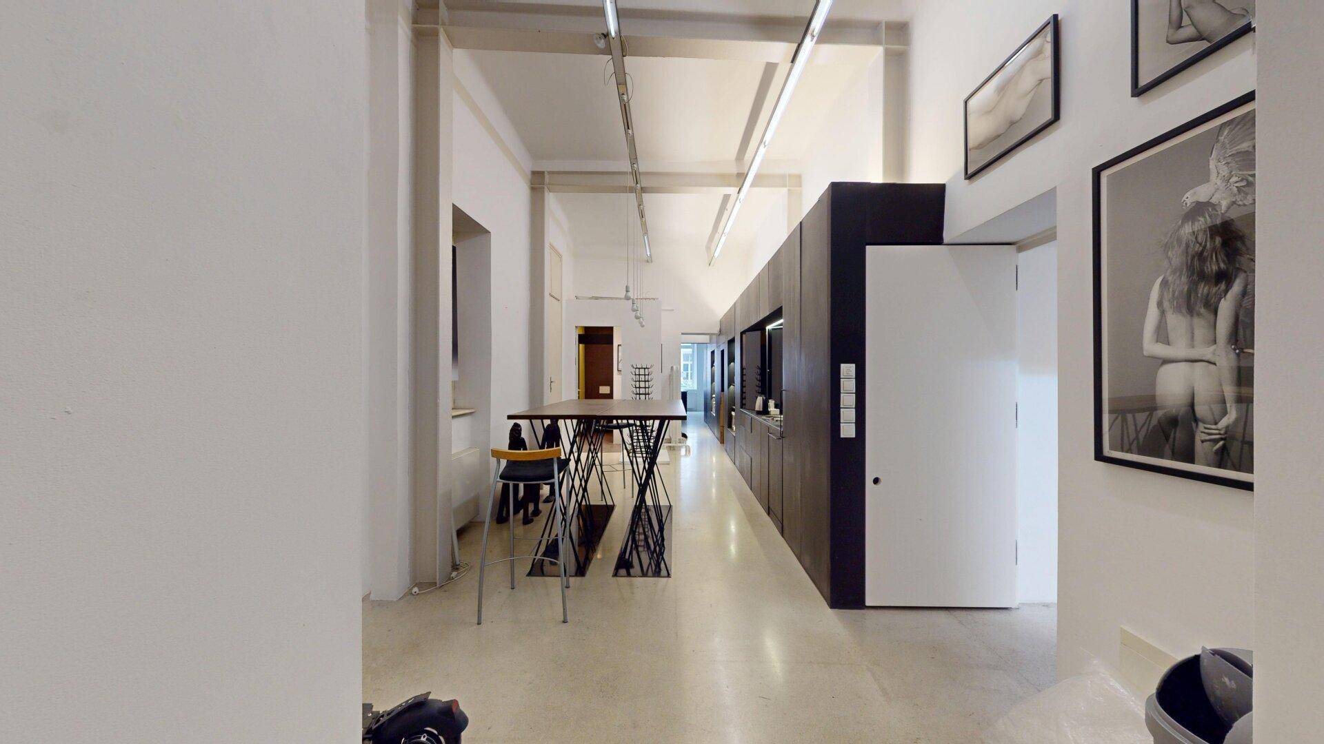 Heller, offener Raum mit Kunstwerken, einem hohen Tisch und einer modernen Küchenzeile im Hintergrund.