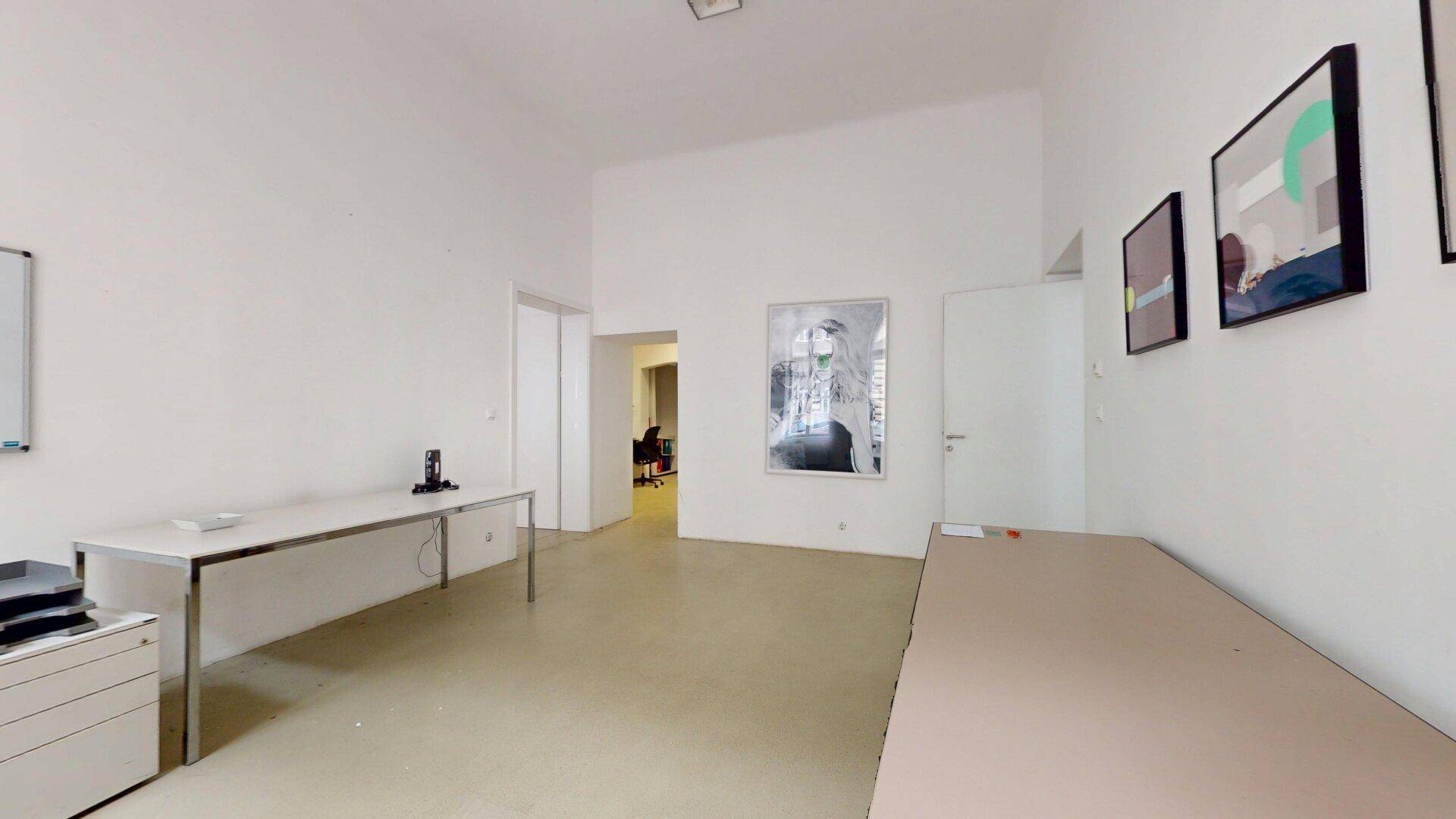Geräumiges Büro mit mehreren Schreibtischen, Kunstwerken an der Wand und einem Durchgang zu einem anderen Raum.