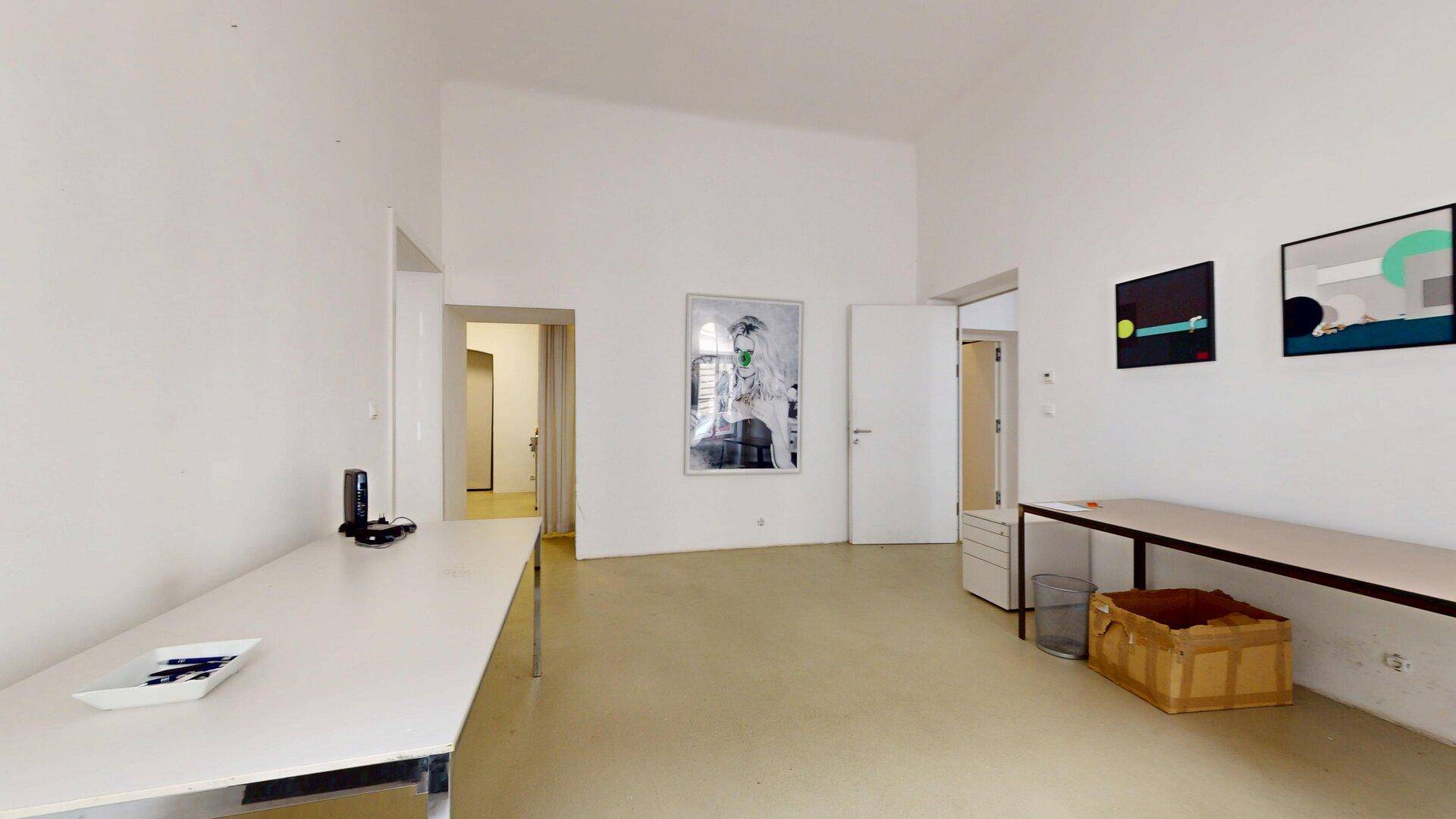 Helles Büro mit Schreibtischen, Kunstwerken und einem offenen Durchgang zu einem weiteren Arbeitsbereich.