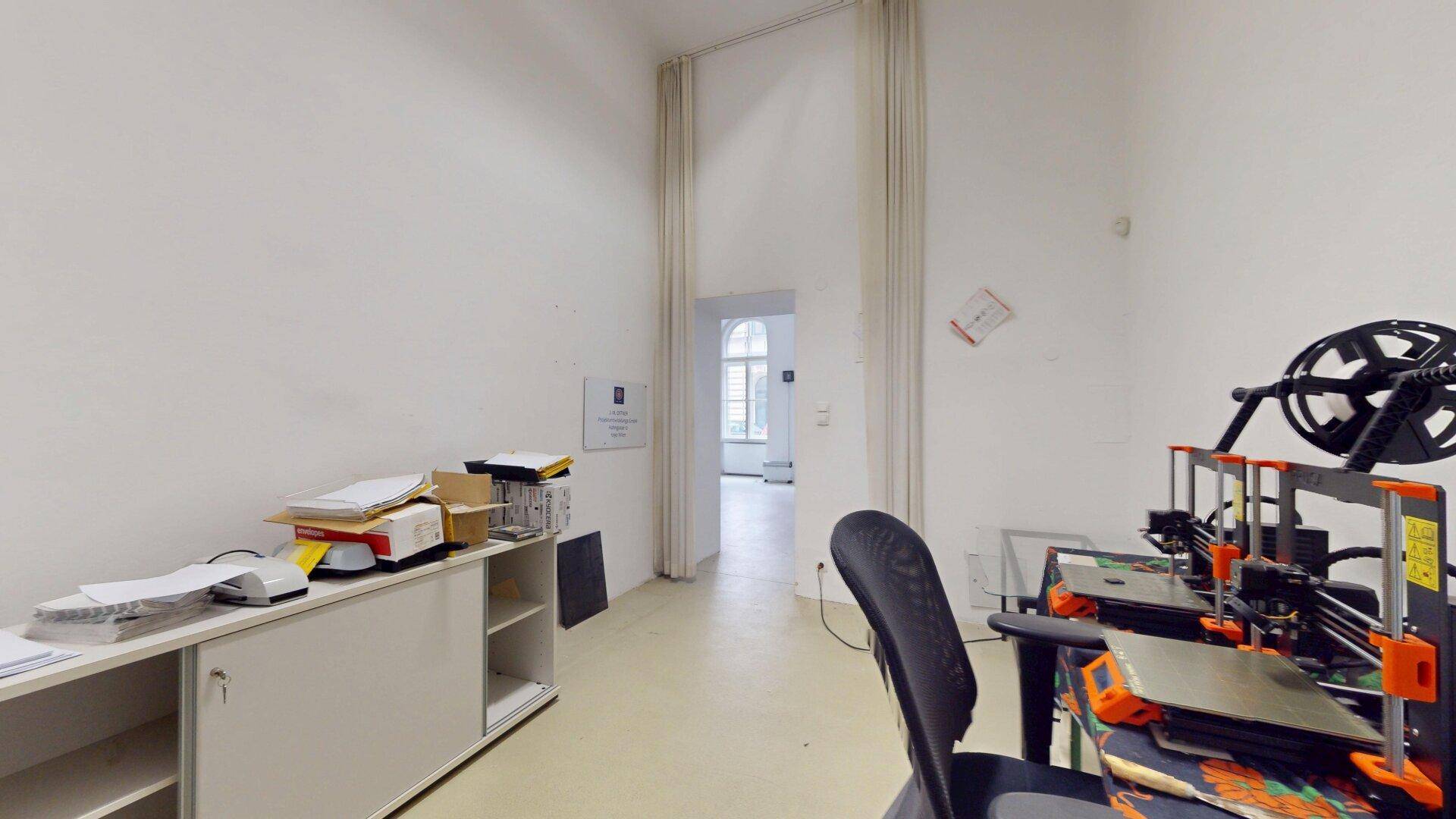 Büroraum mit einem Schreibtisch, Arbeitsgeräten und einem offenen Durchgang zu einem helleren Bereich.