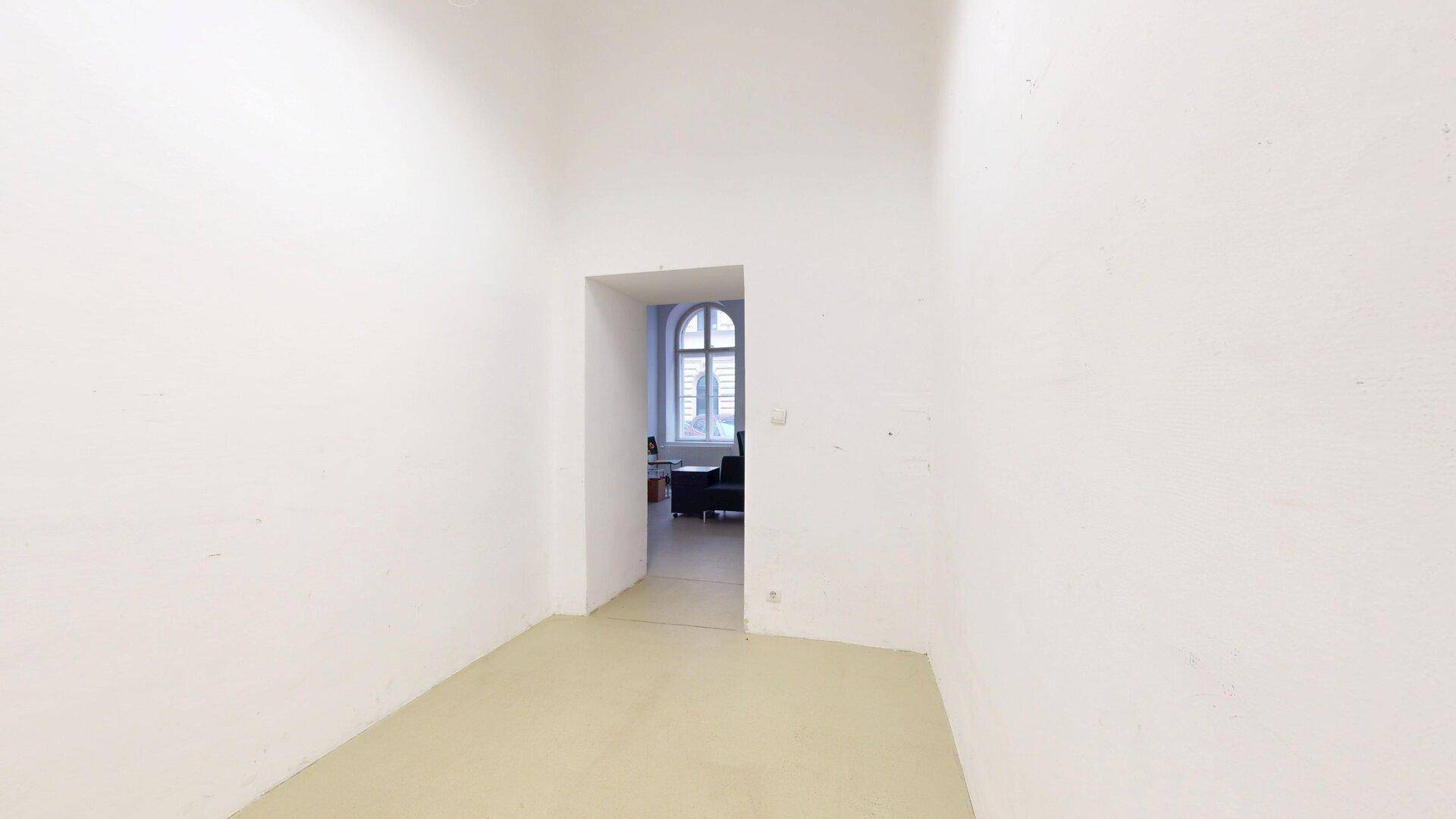 Kleiner, leerer Raum mit weiß gestrichenen Wänden und einem Durchgang zu einem weiteren Bereich.