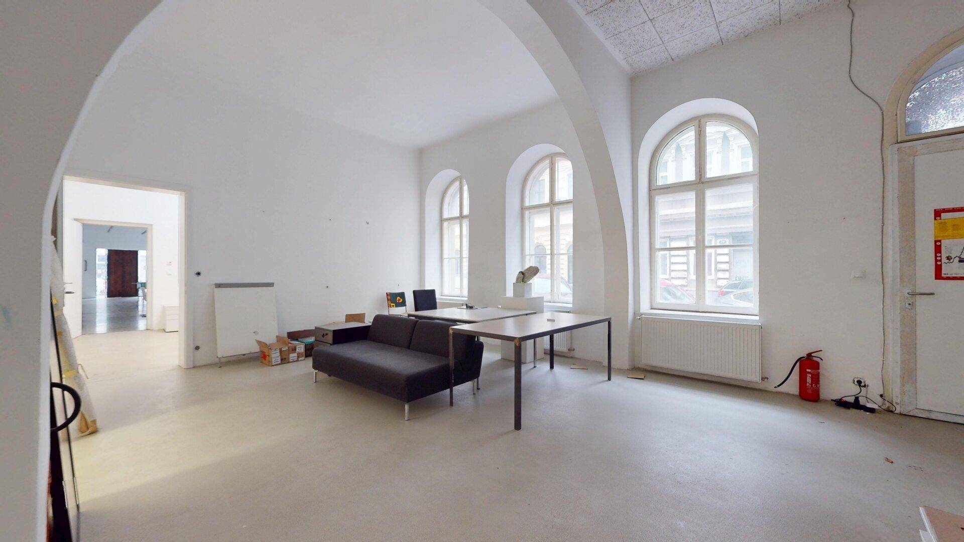 Heller Büroraum mit hohen Decken, großen Rundbogenfenstern und einfacher Büroeinrichtung.