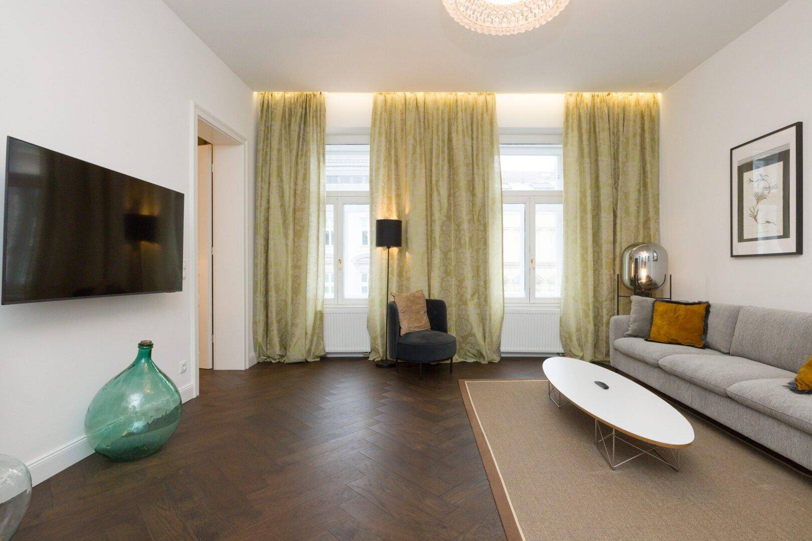 Geräumiges Wohnzimmer mit Wandfernseher, bequemer Sitzgelegenheit und großen Fenstern.