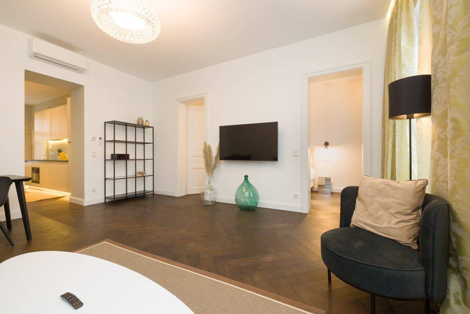 Heller Wohnbereich mit Wandfernseher, Regal und Zugang zu weiteren Räumen.