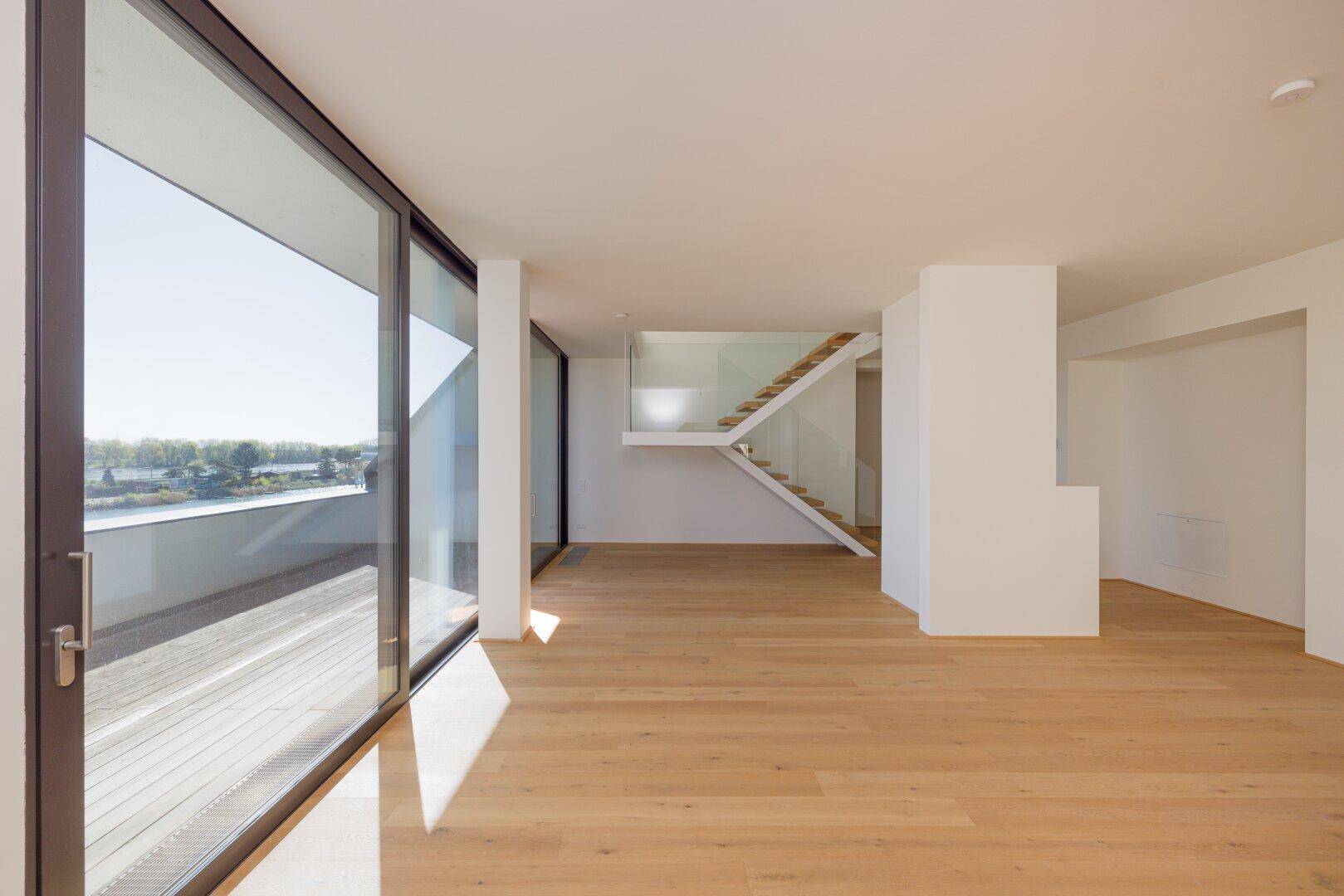 Offener Wohnbereich mit hellem Parkettboden und moderner Treppe mit Glasgeländer.