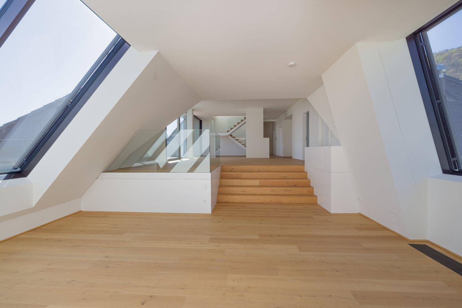 Weitläufiger Dachgeschossraum mit Holzstufen, Glasgeländer und mehreren Schrägfenstern.