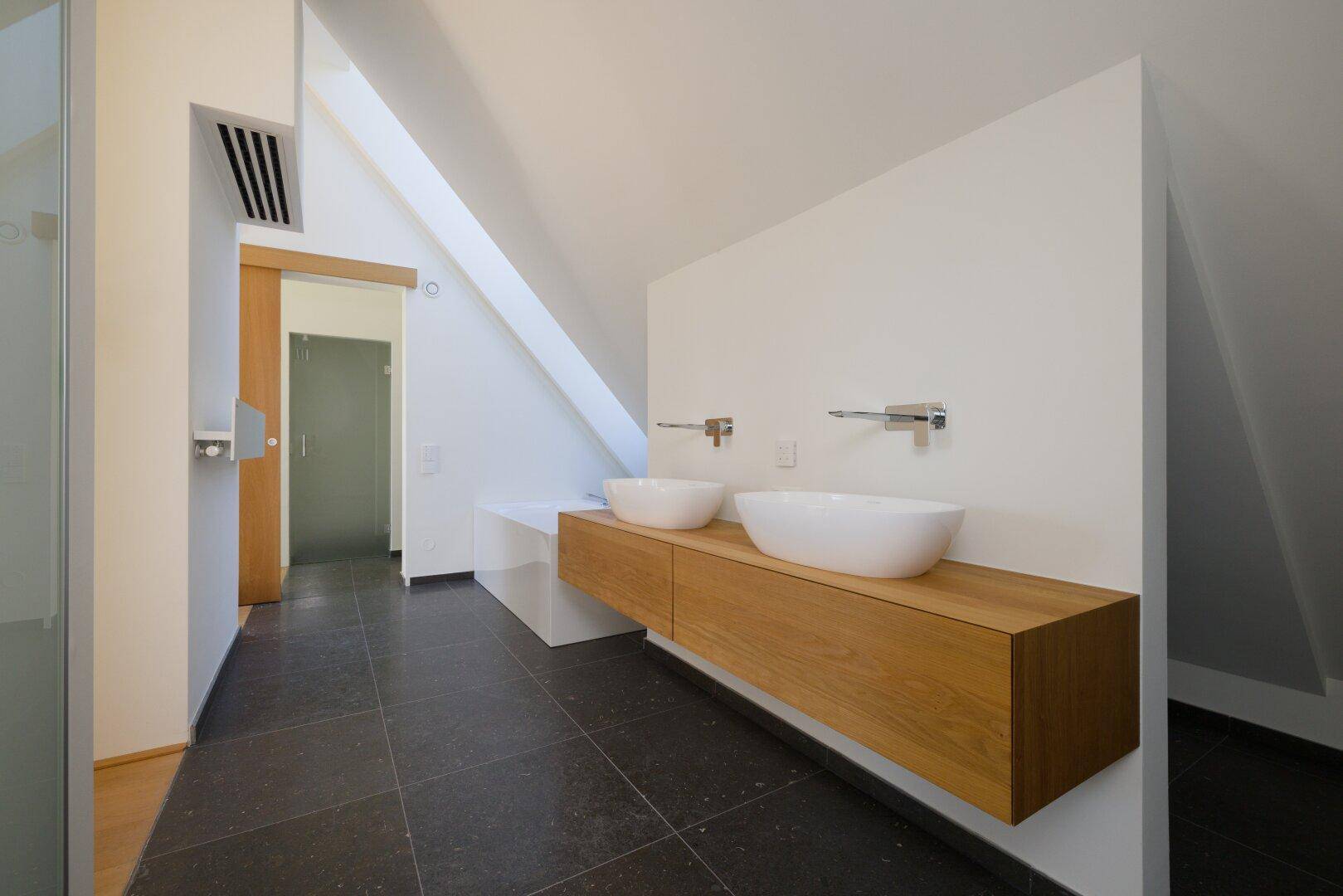 Modernes Badezimmer mit Doppelwaschbecken auf Holzunterschrank und dunklen Bodenfliesen.