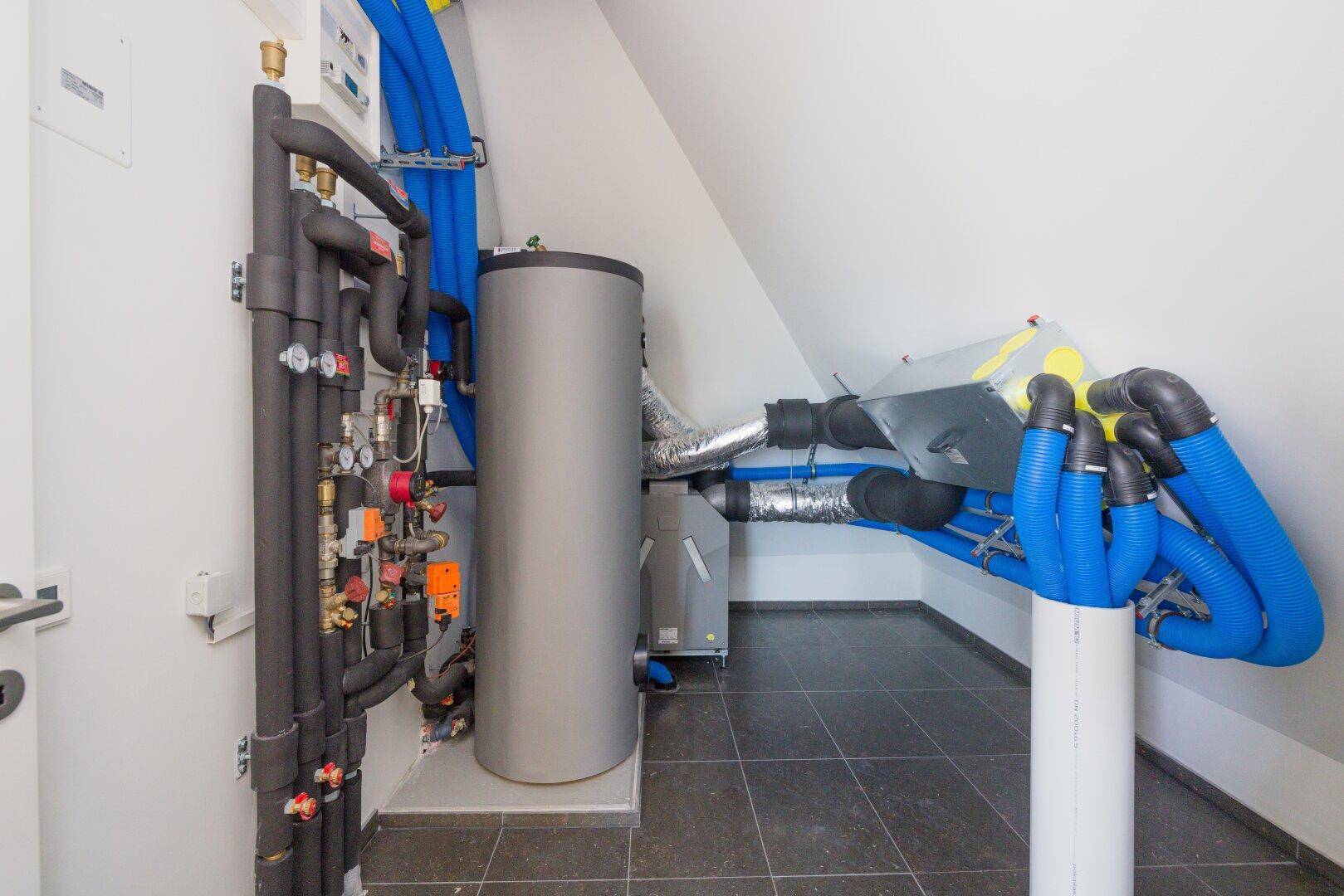 Technikraum mit Heizungsanlage, Warmwasserspeicher und umfangreicher Rohrinstallation unter einer Dachschräge.