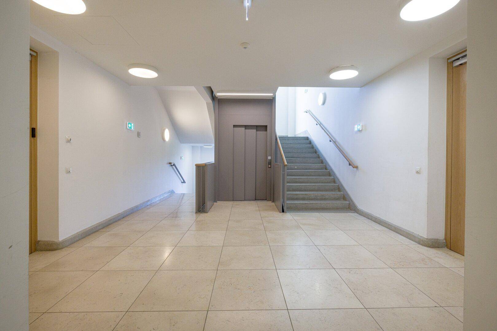 Heller Flur mit Aufzug und Treppenhaus, ausgestattet mit Fliesenboden und modernen Deckenleuchten.