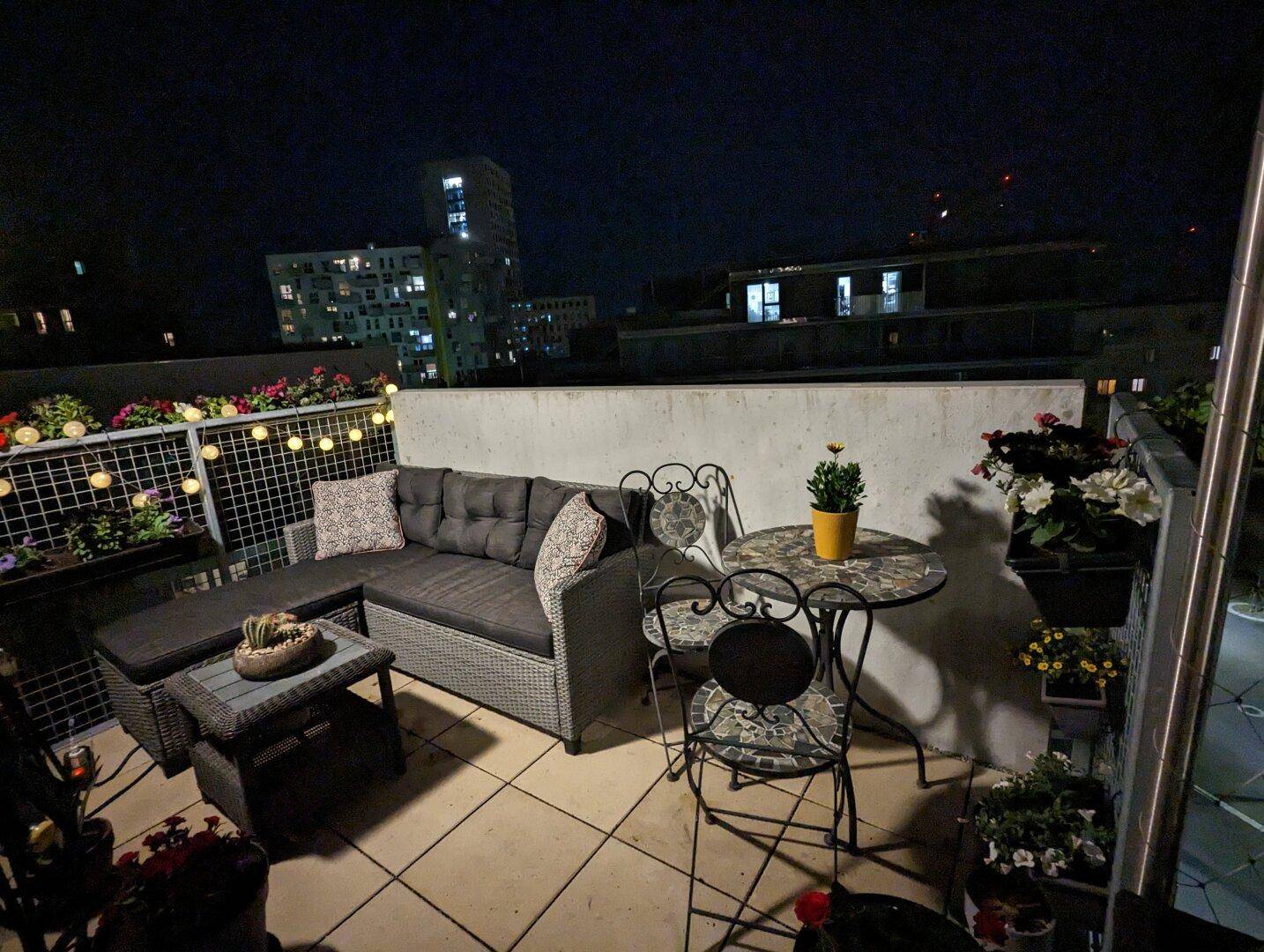 Gemütlicher Balkon bei Nacht mit stimmungsvoller Beleuchtung und Stadtlichtern im Hintergrund.