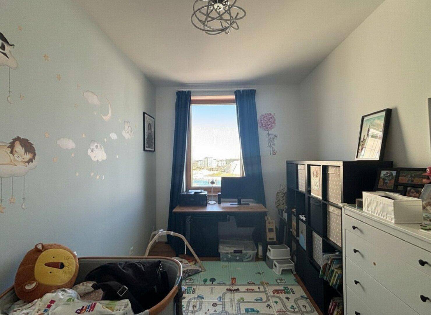 Kinderzimmer mit Schreibtisch, Spielteppich und Fenster mit Tageslicht.