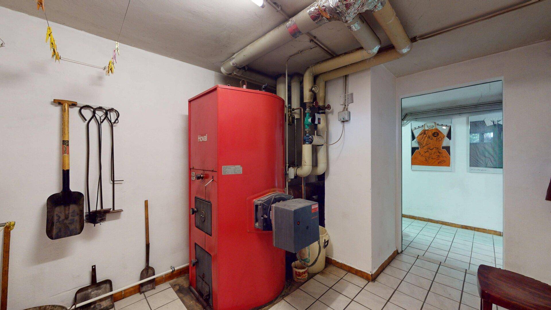 Heizungsraum mit einem großen roten Heizkessel, sichtbaren Rohren und Fliesenboden.