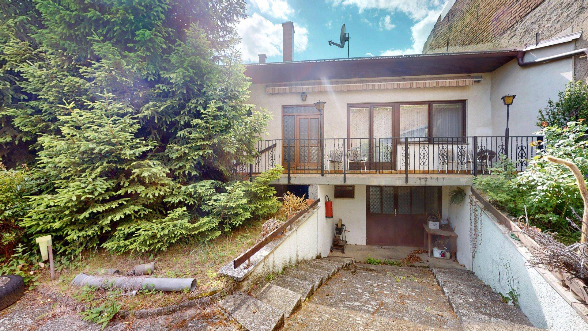 Rückseite des Hauses mit Balkon, Außentreppe zum Keller und umgebender Vegetation.