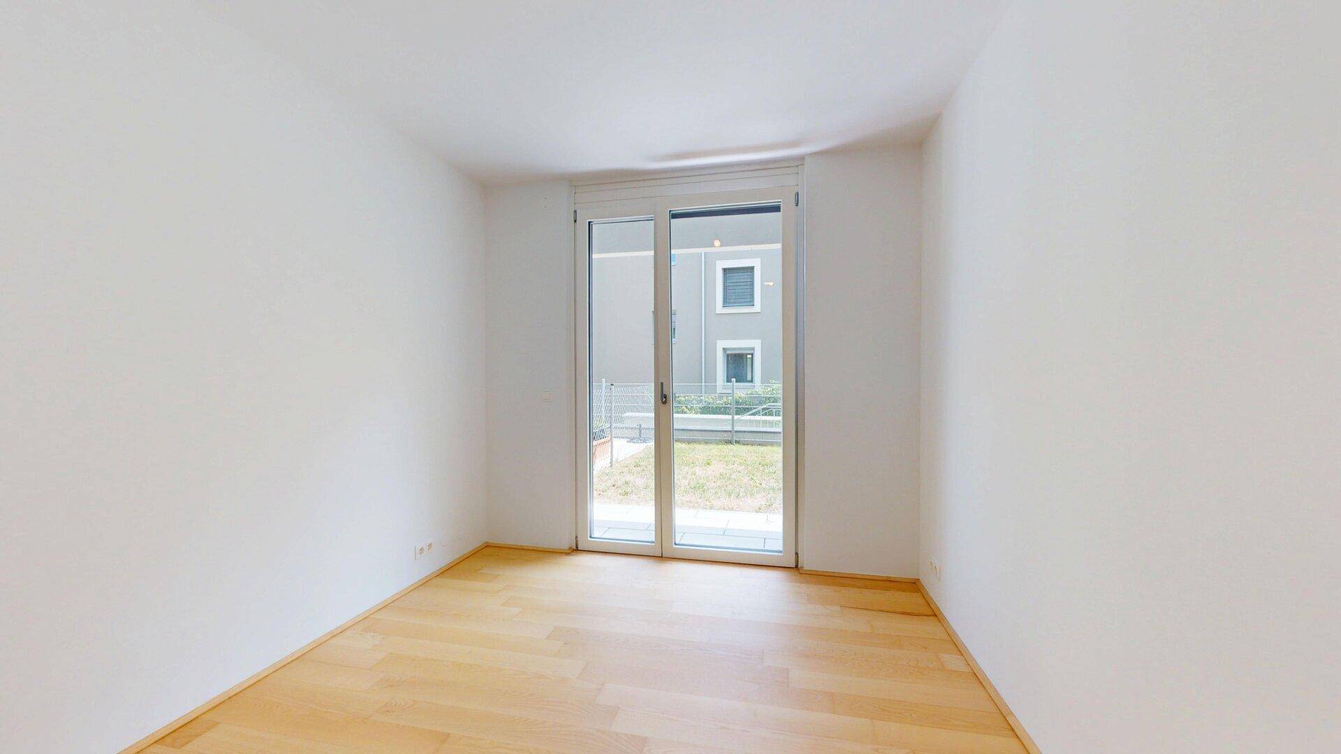 Helles Zimmer mit Holzboden und Zugang zu einem kleinen Außenbereich mit Grünfläche.