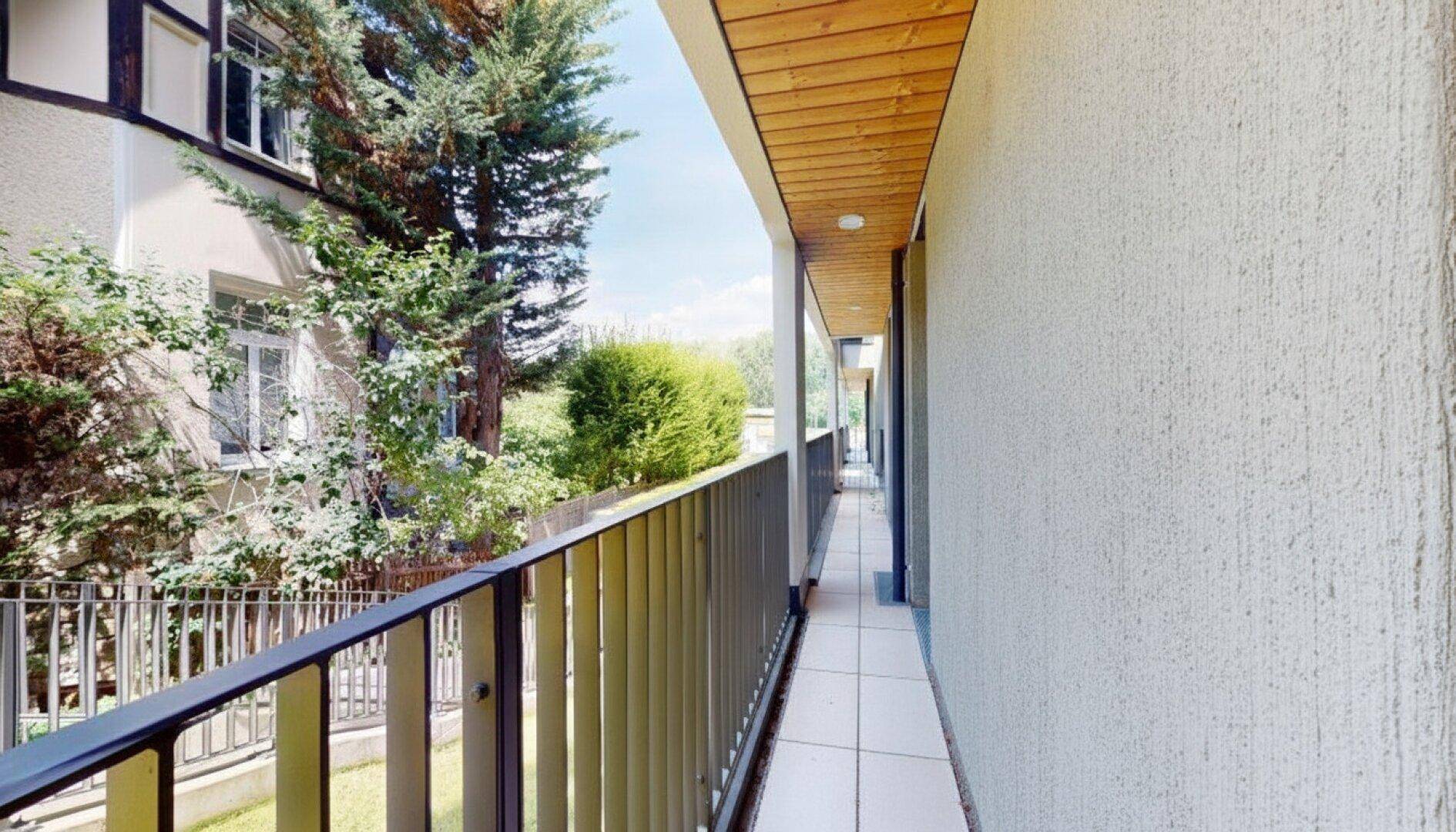Langer, schmaler Balkon mit Holzdach und Fliesenboden, umgeben von viel Grün.
