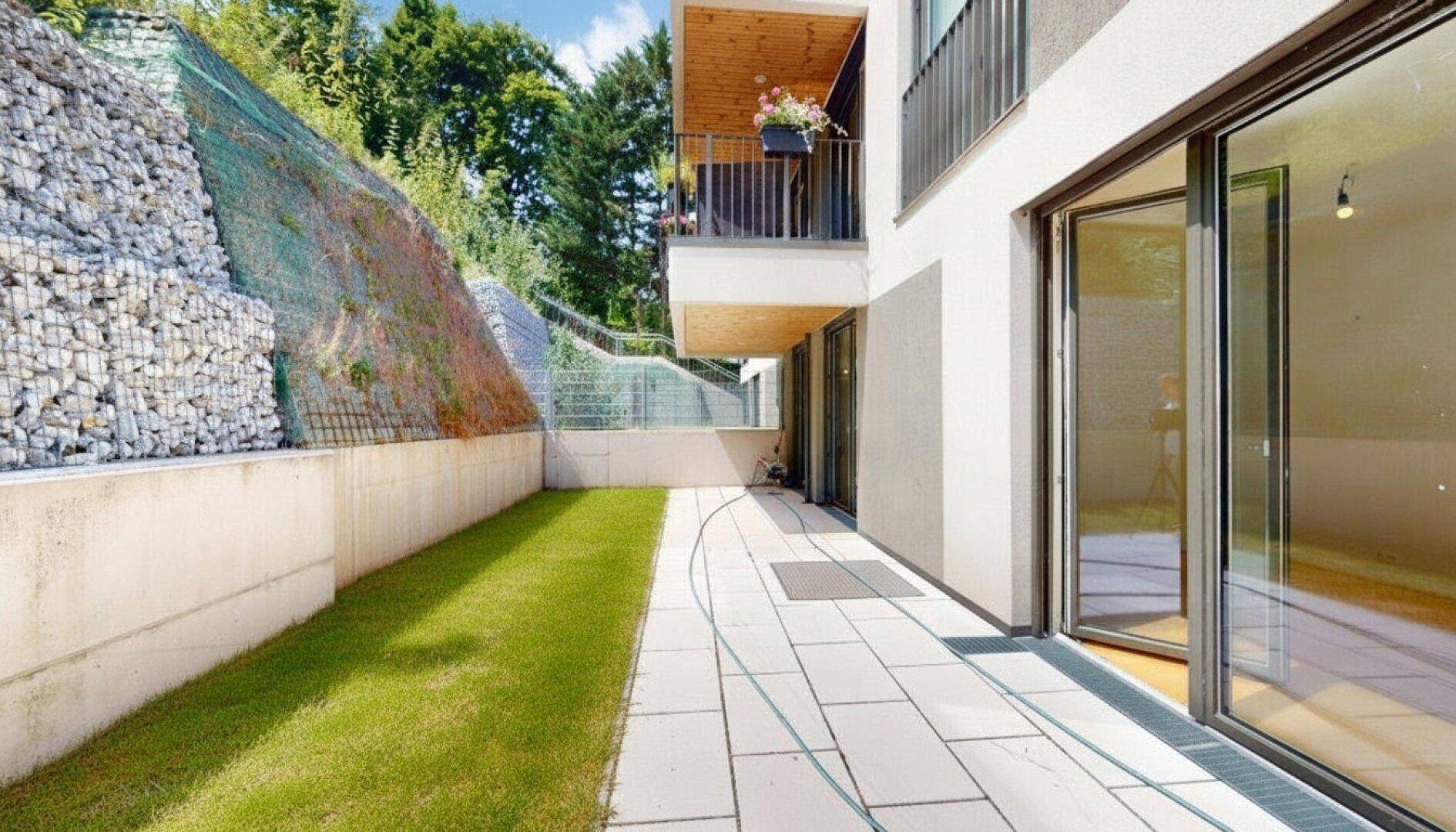 Außenbereich mit gepflasterter Terrasse, Rasenfläche und Gabionenwand neben dem Gebäude.