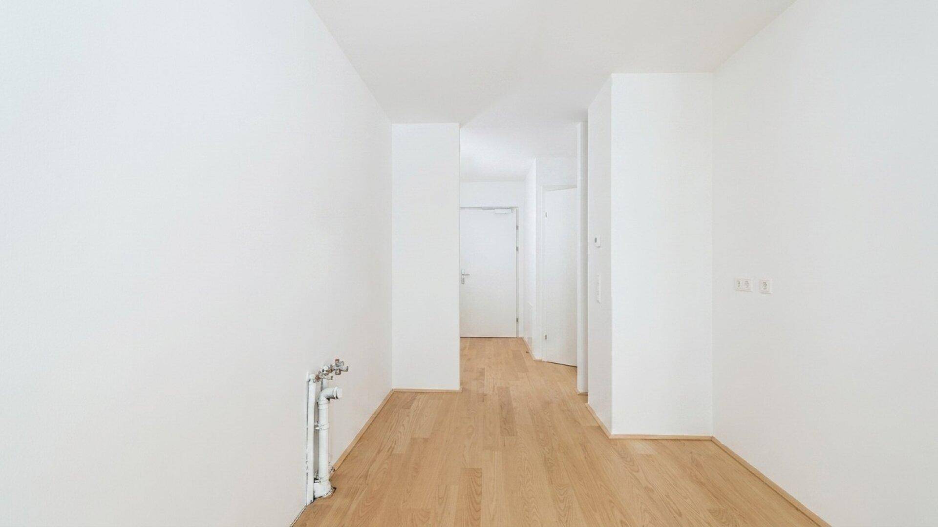 Leerer Raum mit Holzboden und weißen Wänden, vorbereitet für eine Kücheninstallation.