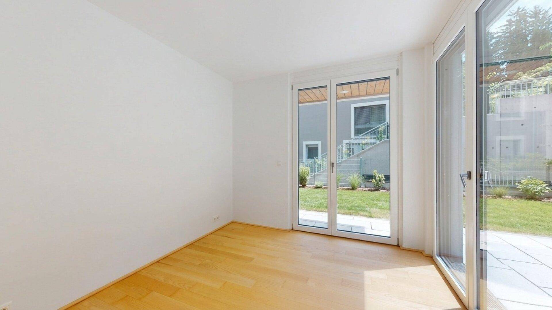 Helles Zimmer mit Holzboden und großen Fenstern, die Zugang zu einem Gartenbereich bieten.